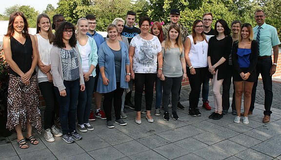 Rund 18 Menschen stehen nebeneinander im Freien und posieren für ein Gruppenfoto. Sie lächeln und tragen sommerliche Alltagskleidung. Im Hintergrund sind Bäume und ein gepflasterter Platz.