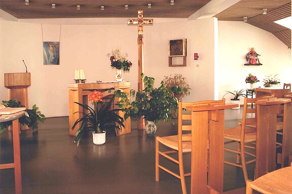 Heller Kapellenraum mit schlichten Holzbänken. Im Zentrum steht ein kleiner Altar mit zwei Kerzen, Blumen und vielen Zimmerpflanzen; dahinter ein großes Kruzifix. Links ein Lesepult, rechts weitere Stühle. Schlichte, moderne Gestaltung, ruhige Andachtsatmosphäre.