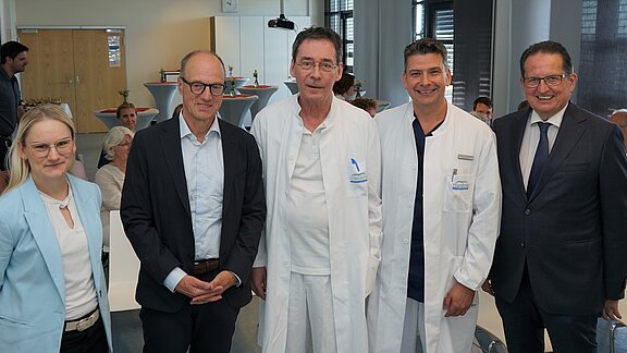 Fünf Personen stehen gemeinsam in einem Raum. Vier Männer tragen helle oder dunkle Anzüge sowie Arztkittel, während eine Frau in einem hellblauen Blazer trägt. Sie wirken freundlich und sind lächelnd. Im Hintergrund sind weitere Personen und einige Tische zu sehen.