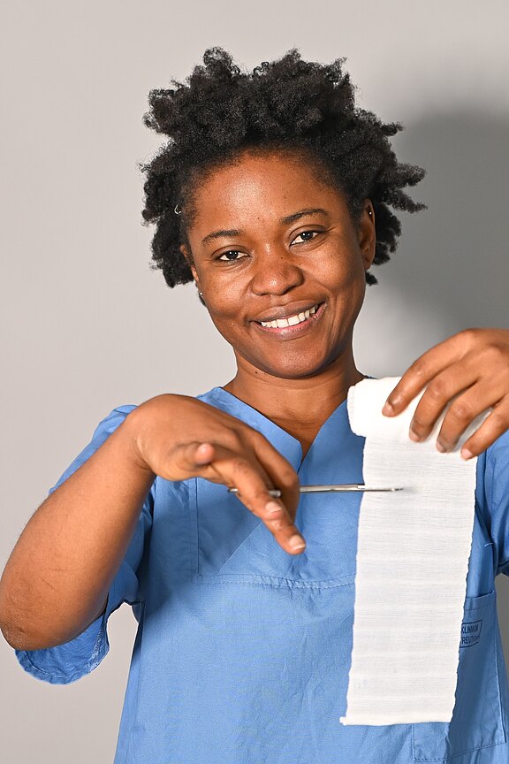 Lächelnde Pflegekraft in blauer OP-Kleidung schneidet mit einer Schere eine weiße Mullbinde ab.