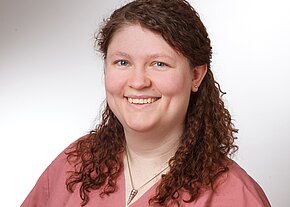 Porträt einer lächelnden Frau mit lockigem braunem Haar. Sie trägt ein rosafarbenes Oberteil und eine Halskette und blickt direkt in die Kamera. Heller, neutraler Hintergrund.