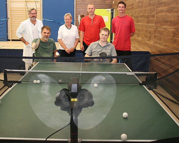 Sechs Erwachsene in einer Sporthalle um eine Tischtennisplatte. Vorn steht ein Trainingsroboter, der Bälle über das Netz schießt. Mehrere Bälle liegen auf der Platte; zwei Personen halten Schläger und wirken bereit zum Üben.
