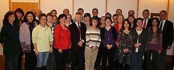 Eine Gruppe von etwa 30 Personen steht lächelnd zusammen in einem Raum, der freundlich beleuchtet ist. Die Teilnehmer sind unterschiedlich gekleidet und scheinen eine gemeinsame Veranstaltung oder ein Treffen zu haben. Die Atmosphäre wirkt positiv und einladend.
