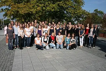 Eine Gruppe von etwa 40 Personen steht im Freien auf einem gepflasterten Platz, umgeben von Bäumen. Die Menschen stehen in mehreren Reihen und lächeln in die Kamera. Viele tragen lässige Kleidung, und die Atmosphäre wirkt freundlich und einladend.