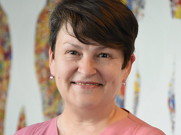 Porträt einer lächelnden Frau mit kurz geschnittenen dunklen Haaren, die einen rosa Kasack trägt. Sie blickt direkt in die Kamera; im Hintergrund sind unscharfe, farbige Formen.