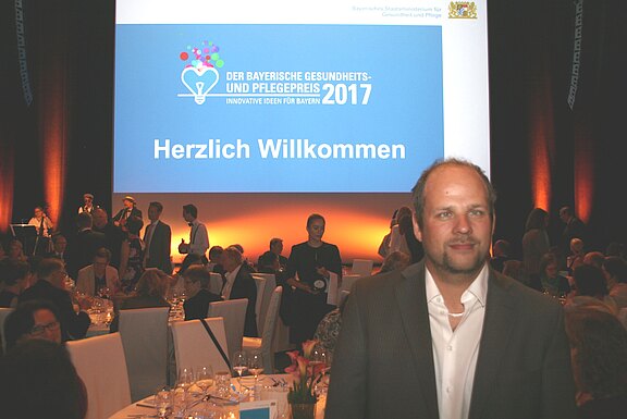 Veranstaltungssaal mit Gästen an Tischen; große Leinwand zeigt: „Der Bayerische Gesundheits- und Pflegepreis 2017 – Innovative Ideen für Bayern. Herzlich Willkommen.“