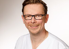 Station 51 Porträt eines lächelnden Mannes mit kurzer Frisur und schwarzer Brille, in weißer medizinischer Kleidung, vor neutralem Hintergrund.