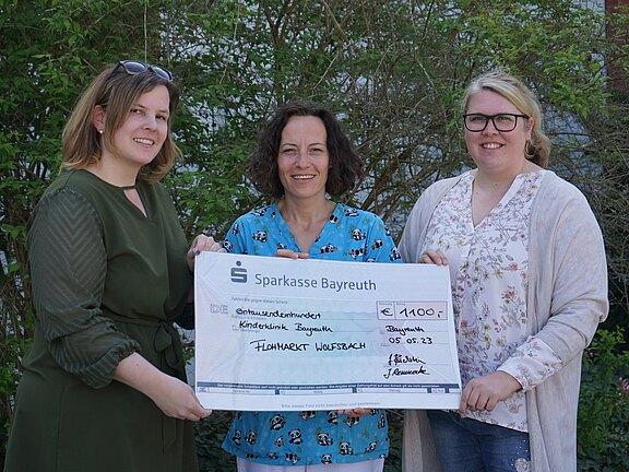 Drei Frauen stehen draußen und halten gemeinsam einen großen Spendenscheck. Der Scheck der Sparkasse Bayreuth über 1.100 Euro ist für die Kinderklinik Bayreuth bestimmt, datiert 05.05.2023, Verwendungszweck: Flohmarkt Wolfsbach. Fröhliche Übergabe.