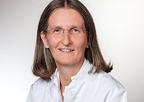 Porträt einer erwachsenen Person mit schulterlangem braun-grauem Haar und Brille, freundlich lächelnd. Sie trägt einen weißen Arztkittel vor neutralem, hellem Hintergrund.