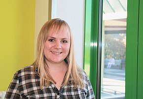 Julia Reichstein Eine junge Frau mit langen blonden Haaren lächelt vor einer hellen Wand und einem Fenster mit grünen Rahmen. Sie trägt ein kariertes Hemd und wirkt freundlich und zugänglich. Im Hintergrund sind unscharfe schemenhafte Figuren zu erkennen.