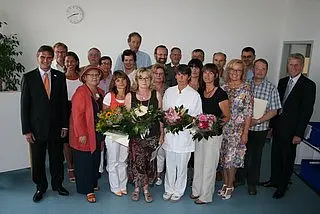 Eine Gruppe von Personen steht in einem Raum zusammen. Einige halten Blumensträuße in den Händen. Es gibt eine Mischung aus Männern und Frauen, die in formeller Kleidung gekleidet sind. Im Hintergrund ist eine Uhr an der Wand sichtbar.
