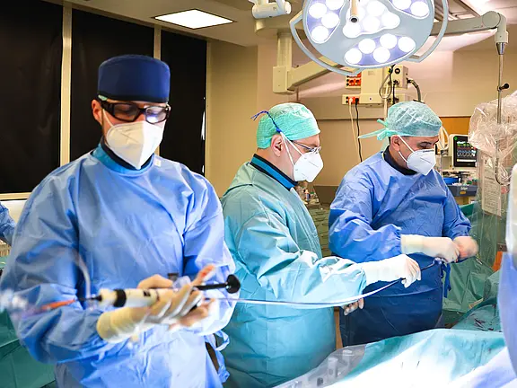 Ärzteteam im Operationssaal in blauen Schutzkitteln und Masken führt einen Eingriff durch. Ein Arzt hält ein katheterähnliches Instrument, während zwei Kolleg:innen am Operationstisch arbeiten. Helles OP-Licht beleuchtet den Bereich; der Patient liegt verdeckt unter Tüchern.