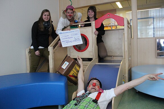 Zwei Clowns in bunten Kostümen präsentieren in einem Kinder-Spielbereich einen großen Spenden-Scheck; einer liegt lachend am Ende einer Rutsche, der andere steht oben an der Spielburg. Zwei weitere Erwachsene lächeln daneben. Fröhliche Szene einer Spendenübergabe für Kinder.