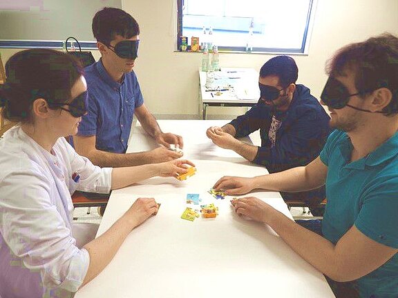 Vier Personen sitzen an einem Tisch, alle tragen Augenbinden. In der Mitte liegen bunte kleine Gegenstände, die sie ertasten und sortieren. Die Szene wirkt wie eine Übung ohne visuelle Hinweise, etwa Sensibilisierung für Blindheit oder Training der taktilen Wahrnehmung.