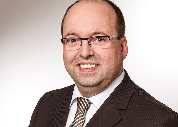 Porträtfoto eines lächelnden Mannes mit Brille und wenig Haar, im dunklen Anzug mit gestreifter Krawatte, vor neutralem, hellen Hintergrund.