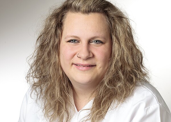 Porträt einer lächelnden Person mit schulterlangen, hellbraunen, leicht lockigen Haaren und Nasenring. Sie trägt ein weißes Oberteil und blickt in die Kamera; der Hintergrund ist hell und neutral.