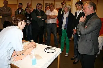 Eine Person in einem weißen Kittel zeigt einer Gruppe von Zuschauern, wie man einen medizinischen Eingriff an einer Puppenpuppe durchführt. Die Zuschauer stehen aufmerksam und beobachten den Prozess, während verschiedene Materialien auf dem Tisch präsentiert werden.