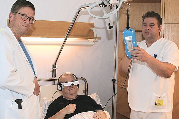 Ein Patient liegt im Krankenhausbett und trägt eine Augenmaske sowie Kopfhörer. Zwei medizinische Fachkräfte stehen neben ihm, einer hält ein Gerät zur Untersuchung in der Hand. Die Szene zeigt eine medizinische Behandlungssituation.