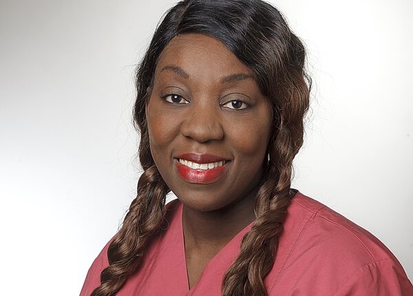 Ein lächelnde Frau mit langen, geflochtenen Haaren trägt ein rotes Oberteil. Ihr Gesicht vermittelt Freundlichkeit und Professionalität. Der Hintergrund ist hell und neutral.