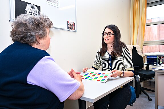 In einem Beratungsraum sitzt eine Fachkraft einer älteren Frau gegenüber. Sie hält ein Blatt mit bunten Quadraten und erklärt eine Aufgabe. Die Szene wirkt wie ein Test- oder Beratungsgespräch.
