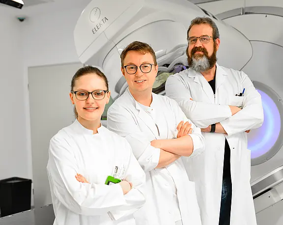 Vor einem großen Strahlentherapiegerät posieren drei medizinische Fachkräfte in weißen Kitteln mit verschränkten Armen und lächeln. Die Szene vermittelt Professionalität in einer modernen Klinik.