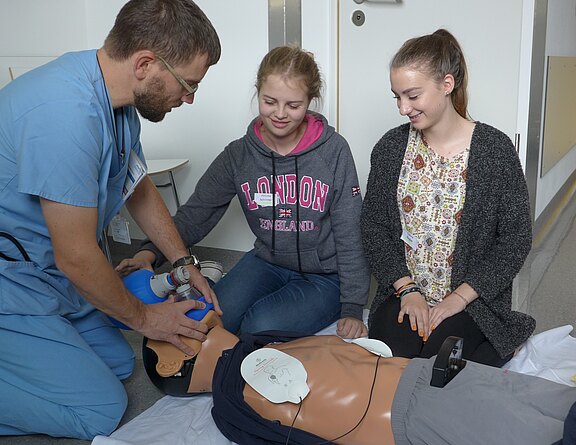 Ein medizinischer Ausbilder demonstriert zwei Jugendlichen an einer Reanimationspuppe die Herz-Lungen-Wiederbelebung. Er nutzt einen Beatmungsbeutel; auf dem Brustkorb liegen Defibrillatorpads. Die Schülerinnen beobachten aus nächster Nähe und üben in einem Schulungsraum.