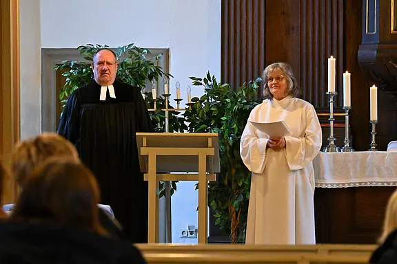 In einer Kirche stehen zwei Geistliche vorne: links ein Mann in schwarzem Talar am Lesepult, rechts eine Frau im weißen Talar mit Blättern in der Hand. Im Hintergrund brennen Kerzen am Altar, im Vordergrund sitzen Gottesdienstbesucher.