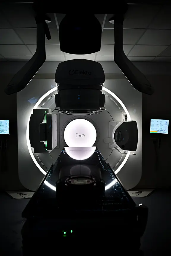 Frontale Ansicht eines modernen Strahlentherapie-Geräts (Linearbeschleuniger) im Klinikraum: ringförmige Gantry mit zentral leuchtendem Kreis „Evo“, davor eine Patientenliege, seitlich Monitore. Die Szenerie ist dunkel, das Gerät dominiert das Bild.
