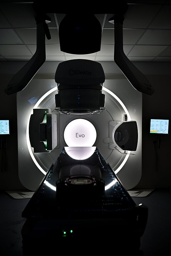 Frontale Ansicht eines modernen Strahlentherapie-Geräts (Linearbeschleuniger) im Klinikraum: ringförmige Gantry mit zentral leuchtendem Kreis „Evo“, davor eine Patientenliege, seitlich Monitore. Die Szenerie ist dunkel, das Gerät dominiert das Bild.
