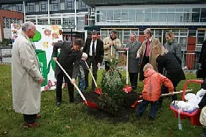 Gruppe von Menschen, darunter Kinder, die gemeinsam einen Baum pflanzen. Einige halten Schaufeln, während sie Erde um den Baum in den Boden verteilen. Im Hintergrund ist ein Gebäude sichtbar. Die Szene vermittelt gemeinschaftliches Engagement für die Natur.