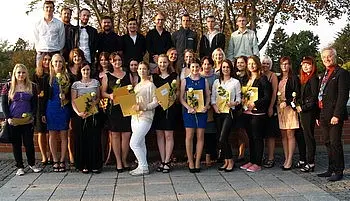 Eine Gruppe von circa 30 Personen steht im Freien zusammen, um einen feierlichen Moment zu teilen. Einige halten Zertifikate in der Hand, während sie Blumen tragen. Die Mehrheit ist in formeller Kleidung gekleidet. Im Hintergrund sind Bäume und eine Abendstimmung sichtbar.