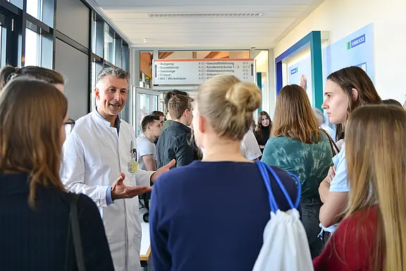 Ein Arzt im weißen Kittel spricht gestikulierend mit einer Gruppe junger Menschen in einem hellen Krankenhausflur; die Gruppe hört zu, als ob es sich um eine Führung oder Informationsveranstaltung handelt.