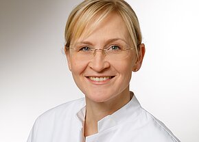 Katharina Wilczek, Dr. rer. nat. Das Bild zeigt eine lächelnde Frau mit blonden Haaren, die eine medizinische Fachjacke trägt. Sie hat eine Brille und steht vor einem hellen, einfarbigen Hintergrund. Ihr Ausdruck vermittelt Freundlichkeit und Professionalität.