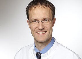 Dr. med. Thomas Prüfer, MHBA Porträt eines lächelnden Mannes mit Brille in weißem Kittel, blauem Hemd und dunkler Krawatte vor neutralem Hintergrund.
