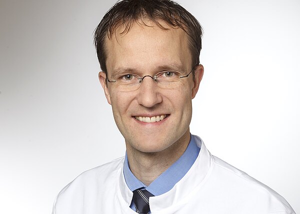 Porträt eines lächelnden Mannes mit Brille in weißem Kittel, blauem Hemd und dunkler Krawatte vor neutralem Hintergrund.