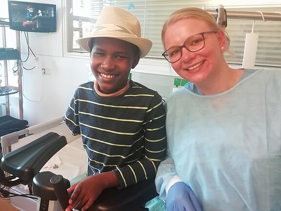 Ein lächelndes Kind mit Strohhut sitzt in einer Klinik am Stuhl. Daneben steht eine freundlich lächelnde medizinische Fachkraft mit Schutzkittel und Handschuhen. Beide blicken in die Kamera; die Szene wirkt warm und unterstützend.