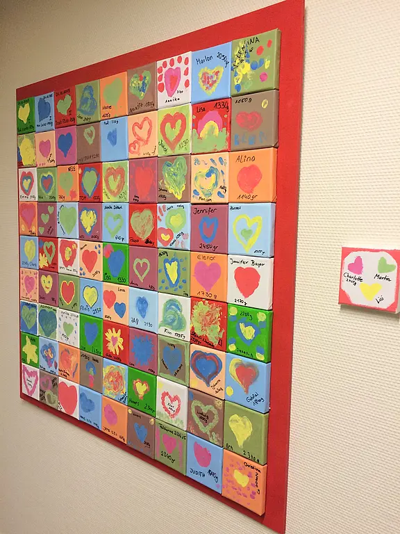 Buntes Wandbild aus vielen kleinen, verschiedenfarbigen Quadraten. In jedem Feld einfache, kindliche Motive, vor allem Herzen, teils mit Vornamen signiert. Insgesamt entsteht ein fröhliches Mosaik.