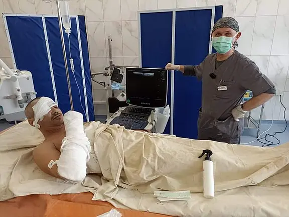 Ein verletzter Patient liegt in einem Krankenhausbett, Augen und rechter Arm stark verbunden. Neben ihm steht eine maskierte Pflegekraft vor einem Ultraschall- bzw. Monitorgerät. Die Szene zeigt medizinische Versorgung nach einer Verletzung oder Operation in einem Klinikraum.