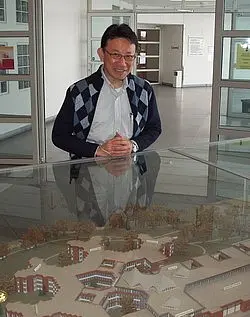Eine Person steht vor einem Modell eines Gebäudekomplexes, das auf einem Tisch liegt. Die Person trägt einen grauen Pullover und schaut freundlich. Das Modell zeigt mehrere Gebäude in einer umliegenden Landschaft.