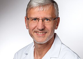 Stefan Huber Porträt eines lächelnden Mannes mit grauen Haaren und Brille, im weißen Kittel, vor neutralem Hintergrund. Kopf und Schultern sind sichtbar.