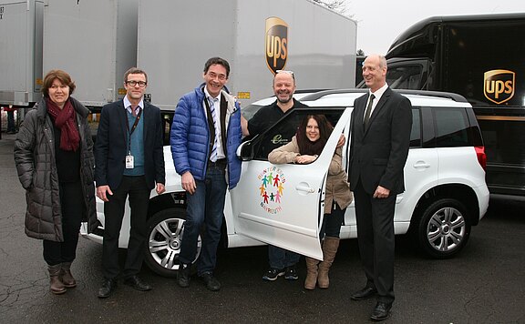 Sechs Erwachsene posieren neben einem weißen SUV; eine Frau sitzt an der geöffneten Beifahrertür. Auf der Tür ein buntes Logo mit Figuren. Im Hintergrund stehen braune Lieferfahrzeuge mit UPS-Logo. Atmosphäre eines Präsentationsfotos.