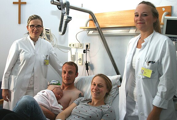 Krankenhauszimmer nach der Geburt: Vater hält ein eingewickeltes Neugeborenes auf der nackten Brust, die Mutter liegt daneben lächelnd im Bett. Zwei Ärztinnen oder Pflegekräfte stehen daneben und schauen freundlich.