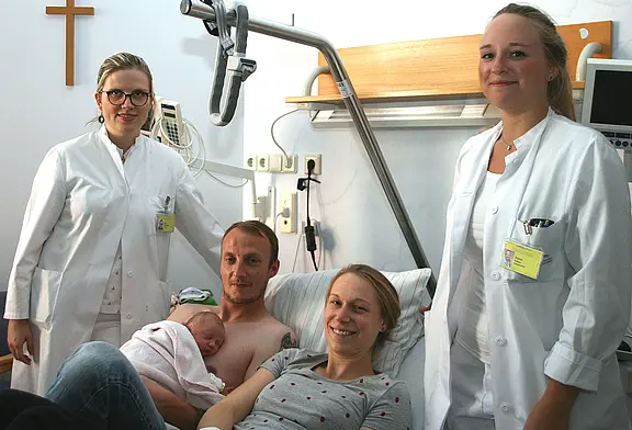 Krankenhauszimmer nach der Geburt: Vater hält ein eingewickeltes Neugeborenes auf der nackten Brust, die Mutter liegt daneben lächelnd im Bett. Zwei Ärztinnen oder Pflegekräfte stehen daneben und schauen freundlich.