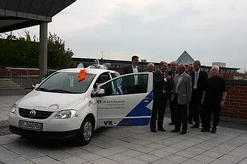 Eine Gruppe von Männern steht um ein weißes Auto herum, das mit einem Band dekoriert ist. Das Auto ist Teil einer Veranstaltung, wahrscheinlich eine Fahrzeugübergabe oder -präsentation. Hinter ihnen ist ein moderner Gebäudekomplex sichtbar.
