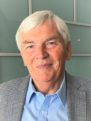 Werner Keller Porträt eines älteren Mannes mit kurzen grauen Haaren und freundlichem Ausdruck. Er trägt ein blaues Hemd und graues Sakko vor neutralem Hintergrund.