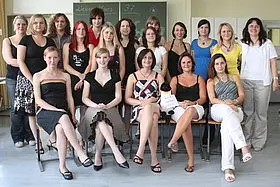 Eine Gruppe von 24 Frauen steht und sitzt in einem Raum. Sie sind in verschiedenen Outfits gekleidet und lächeln in die Kamera. Im Hintergrund sind Tafel und Fenster zu erkennen. Die Atmosphäre wirkt freundlich und gemeinschaftlich.