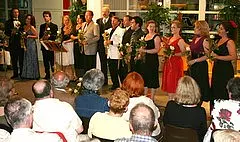 Eine Gruppe von Darstellern steht auf einer Bühne und hält Blumengestecke in den Händen. Sie sind festlich gekleidet und lächeln. Im Vordergrund sind die Köpfe einer Gästen zu sehen, die auf die Darbietung blicken. Die Atmosphäre ist feierlich und ausgelassen.