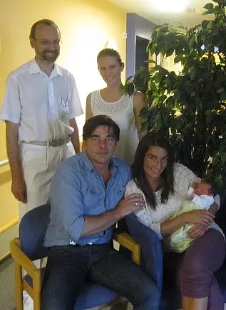 Eine Gruppe von fünf Personen steht und sitzt in einem hellen Raum. Eine Frau hält ein Baby in ihren Armen. Der Mann neben ihr sieht lächelnd in die Kamera. Im Hintergrund ist ein bepflanzter Bereich sichtbar. Die Atmosphäre wirkt freundlich und familiär.