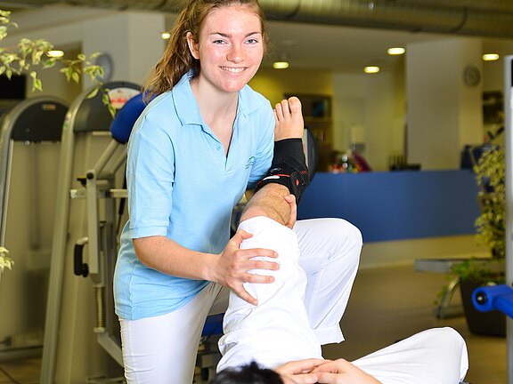 Physiotherapeutin behandelt das Bein eines liegenden Patienten: Sie hebt das bandagierte Sprunggelenk und führt eine Mobilisationsübung in einer Reha-Praxis durch.
