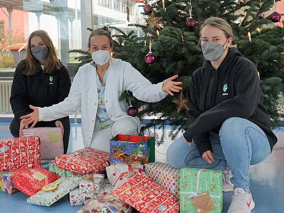 Drei maskierte Personen – eine im weißen Kittel – sitzen vor einem geschmückten Weihnachtsbaum. Vor ihnen liegen viele bunt verpackte Geschenke. Die Szene wirkt wie eine Geschenkaktion oder Spendenübergabe in einem öffentlichen Gebäude.